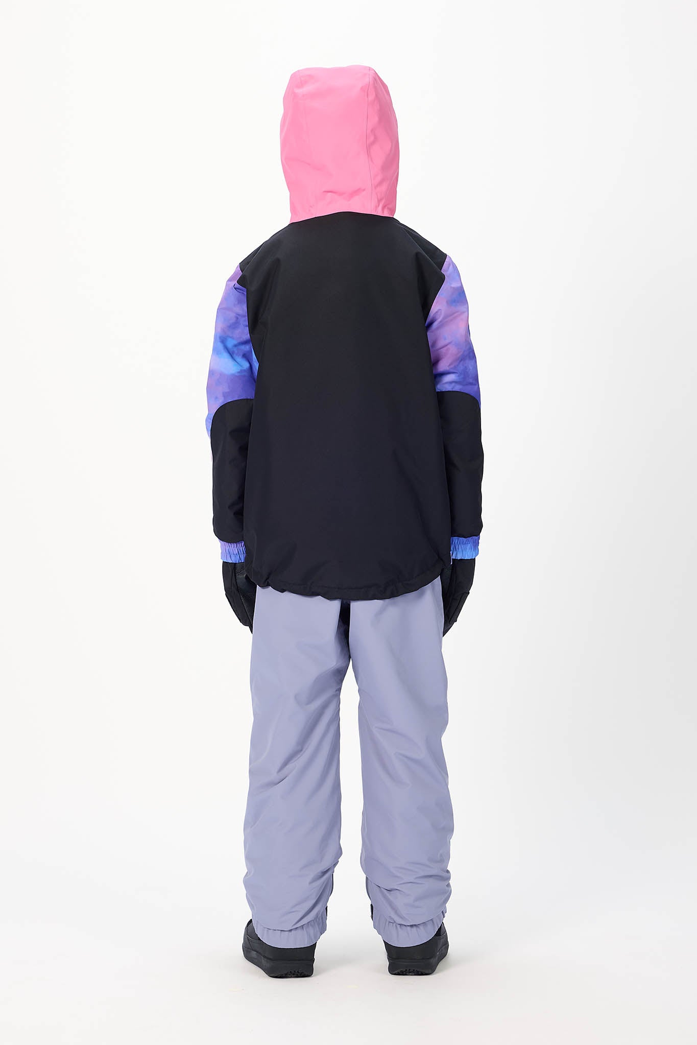 A child shown from the back wearing a color-blocked winter jacket with a bright pink hood, black body, and purple-blue patterned sleeves, paired with light lavender snow pants, against a plain white studio background.